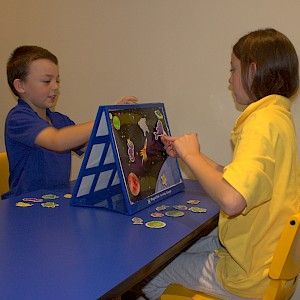 two children sitting at a blue table playing with a board game on the easel