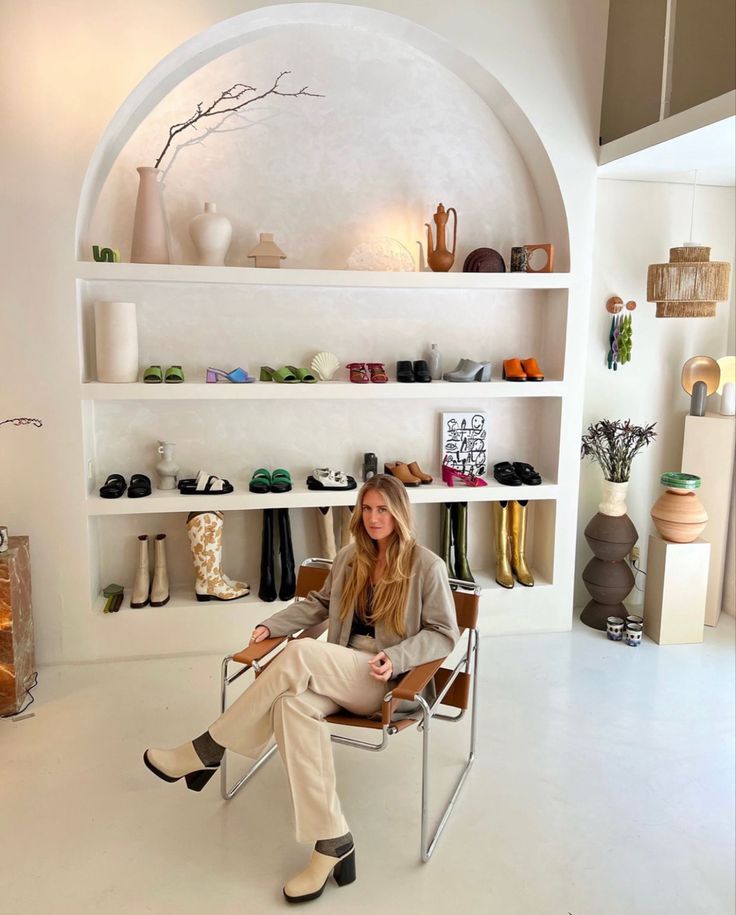 a woman sitting on a chair in front of a shelf with shoes and vases