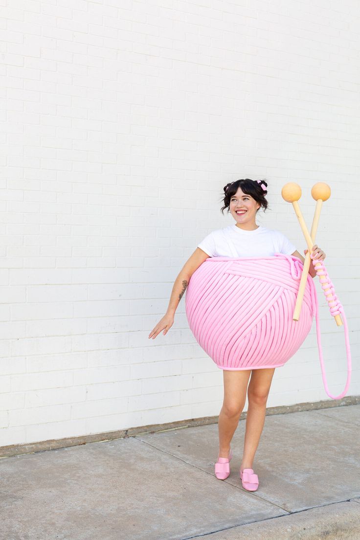 a woman in a pink skirt holding two knitting needles and a large ball of yarn