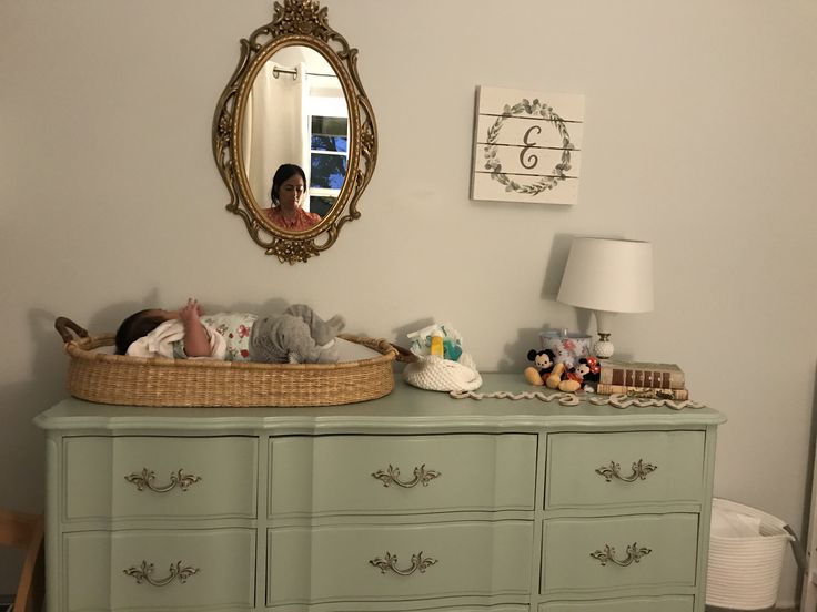 a baby laying in a basket on top of a green dresser next to a mirror