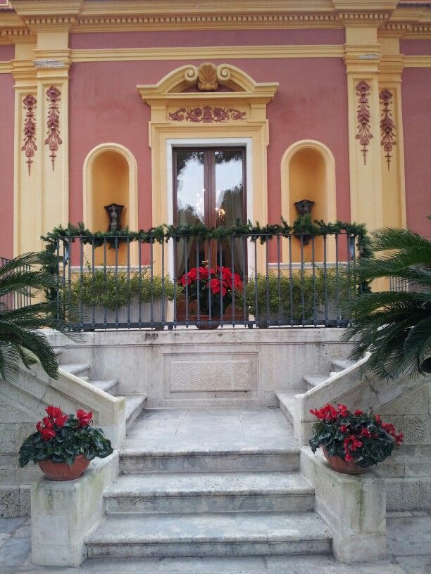 two planters with red flowers are on the steps to a large building that is painted pink and yellow