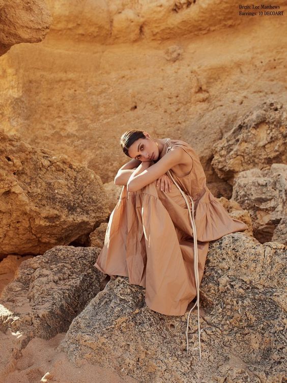 a woman sitting on top of a rock next to a large stone wall and wearing a brown dress