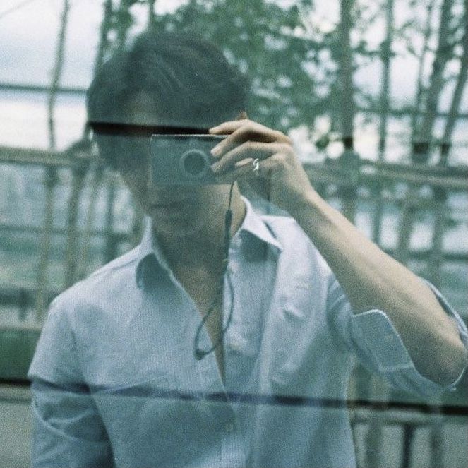 a man taking a selfie in front of a glass wall with trees behind him