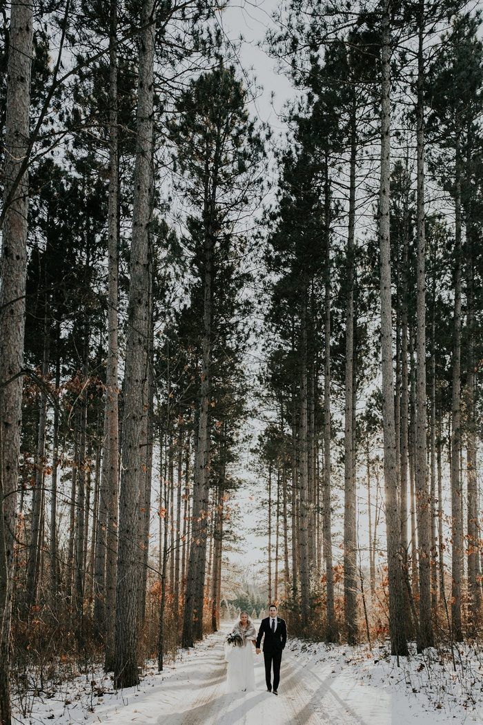 a bride and groom are walking through the snow in front of tall pine trees on their wedding day