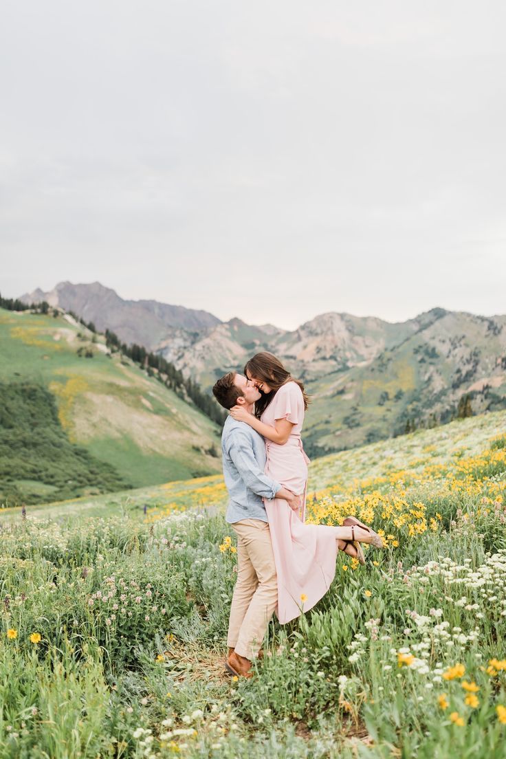 an engaged couple kissing in a field with wildflowers and mountains in the background