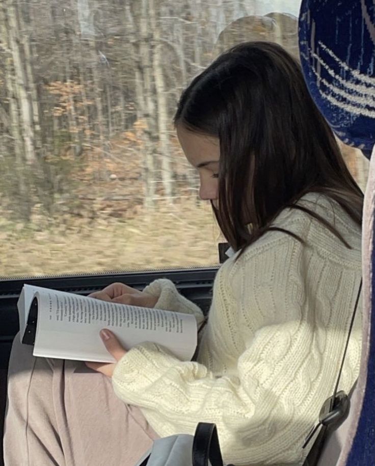 a woman sitting in a seat on a bus reading a book and looking out the window