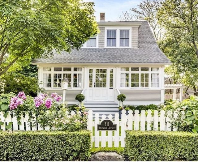 a white picket fence in front of a house with flowers and bushes around it,