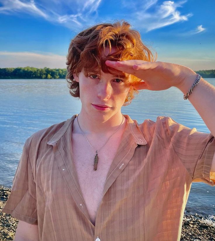 a young man holding his hand up to his head while standing on the shore of a lake