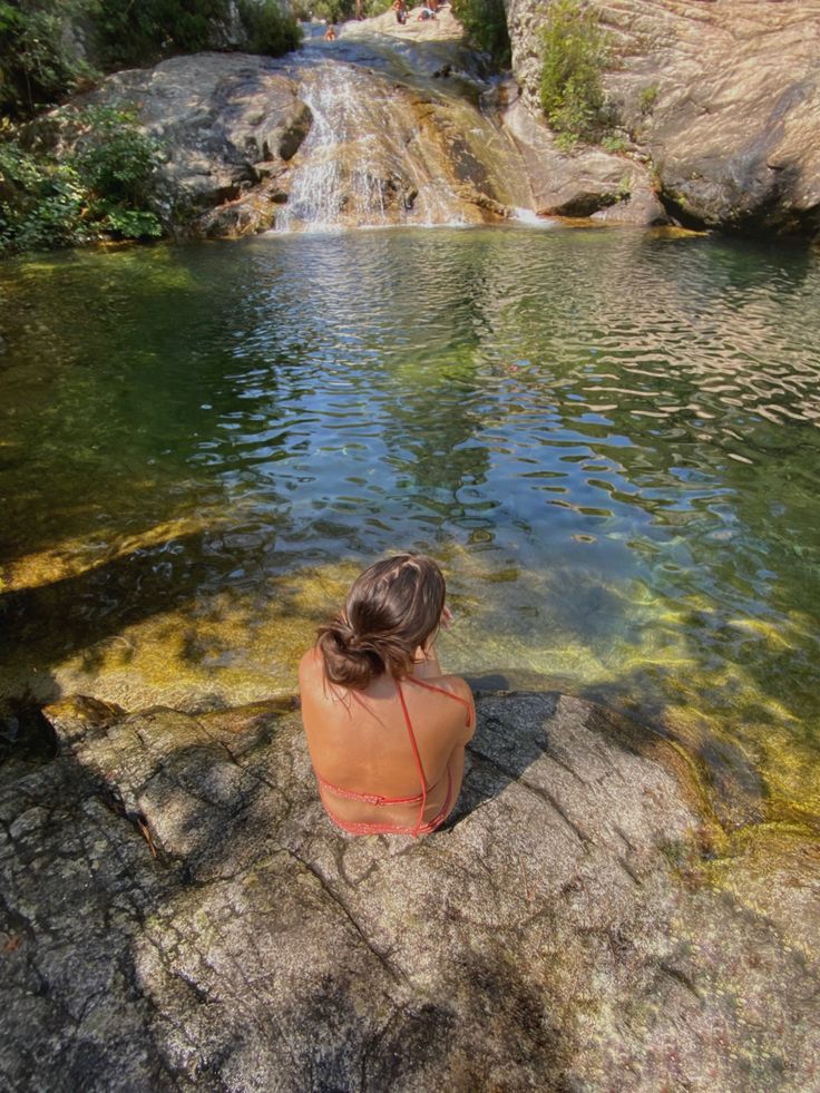 a woman sitting on top of a rock next to a body of water with a waterfall in the background