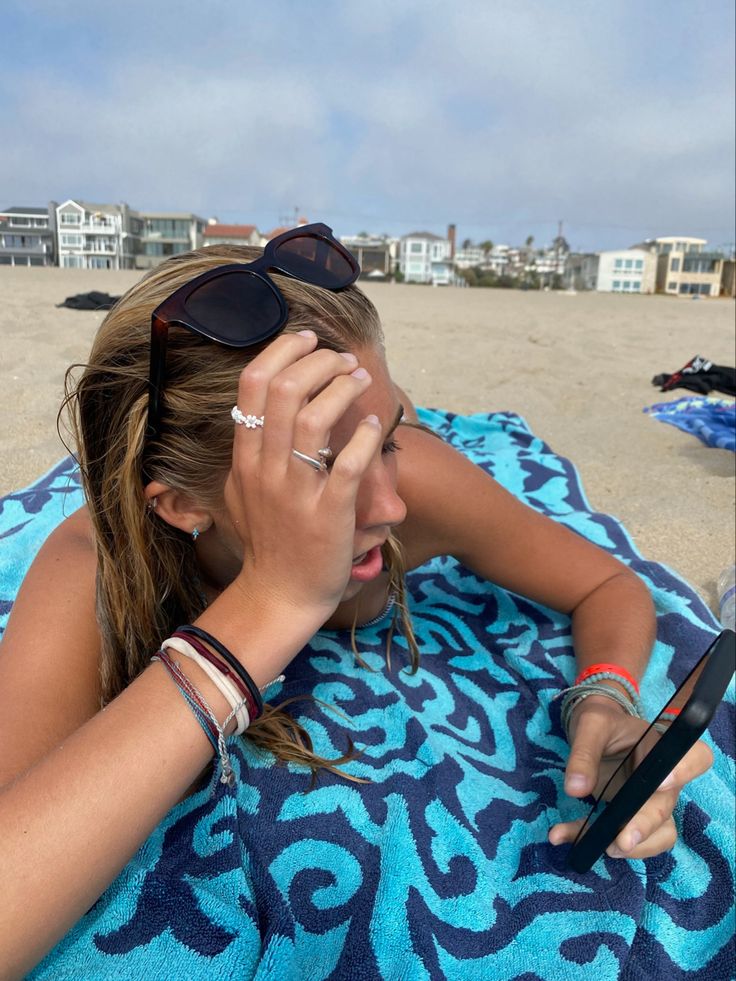 a woman laying on top of a blue towel next to the ocean holding a cell phone