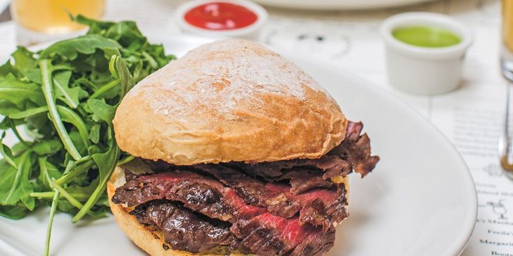 a steak sandwich on a white plate with greens and orange juices in the background