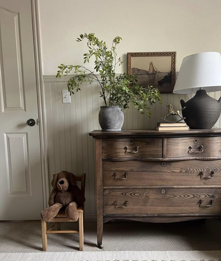 a teddy bear sitting on a chair in front of a dresser with a lamp and potted plant