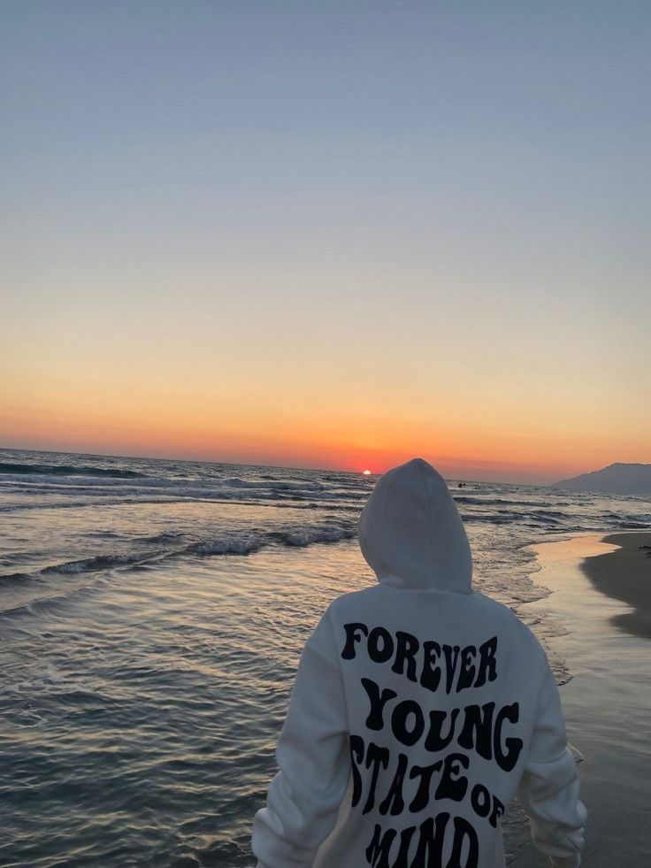 a person standing on the beach at sunset with their back to the camera, wearing a hoodie that says forever young state of mind