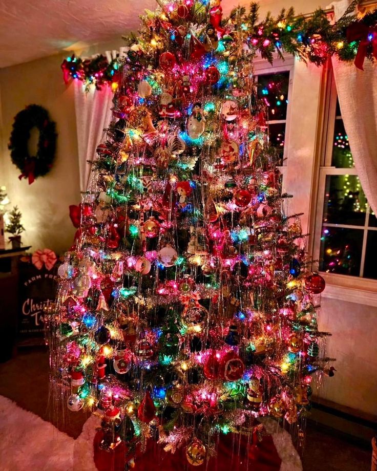 a brightly colored christmas tree in a living room next to a window with lights on it