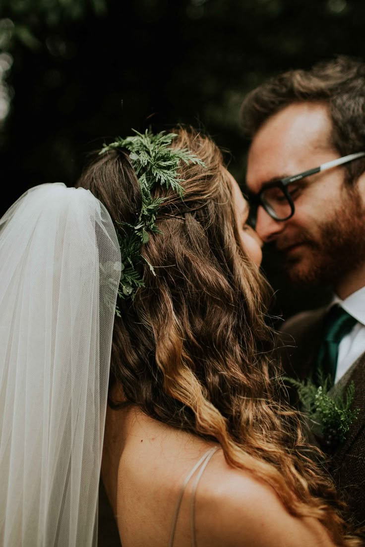 a bride and groom kissing in front of trees at their outdoor wedding ceremony with greenery on the veil