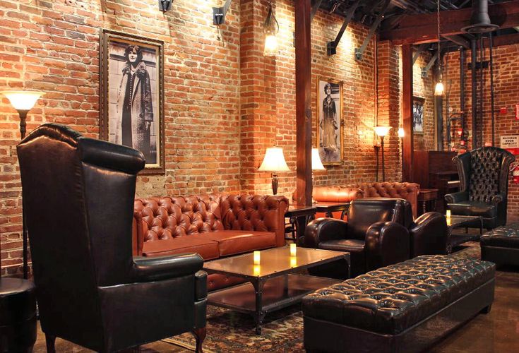 a living room filled with lots of brown furniture and brick walls, along with two lamps on either side of the couches