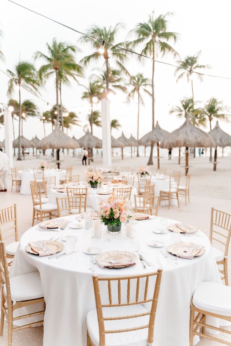 a table set up with white linens and gold chairs for an outdoor wedding reception on the beach