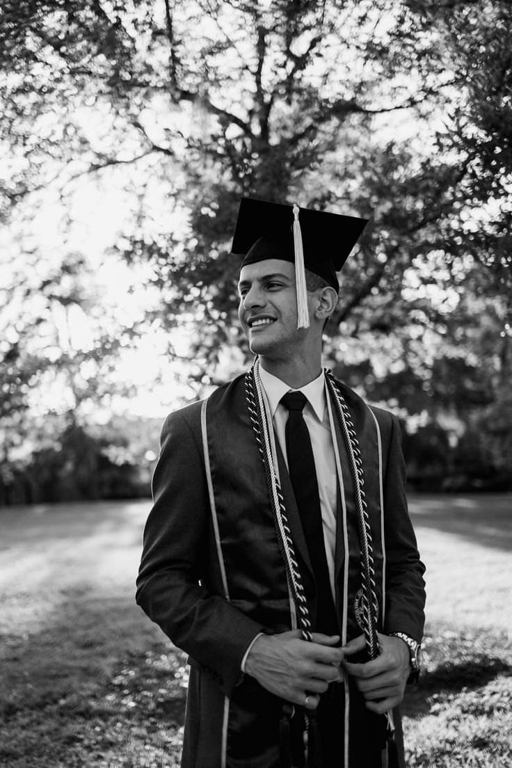 a man in a graduation cap and gown standing under a tree with his hands in his pockets