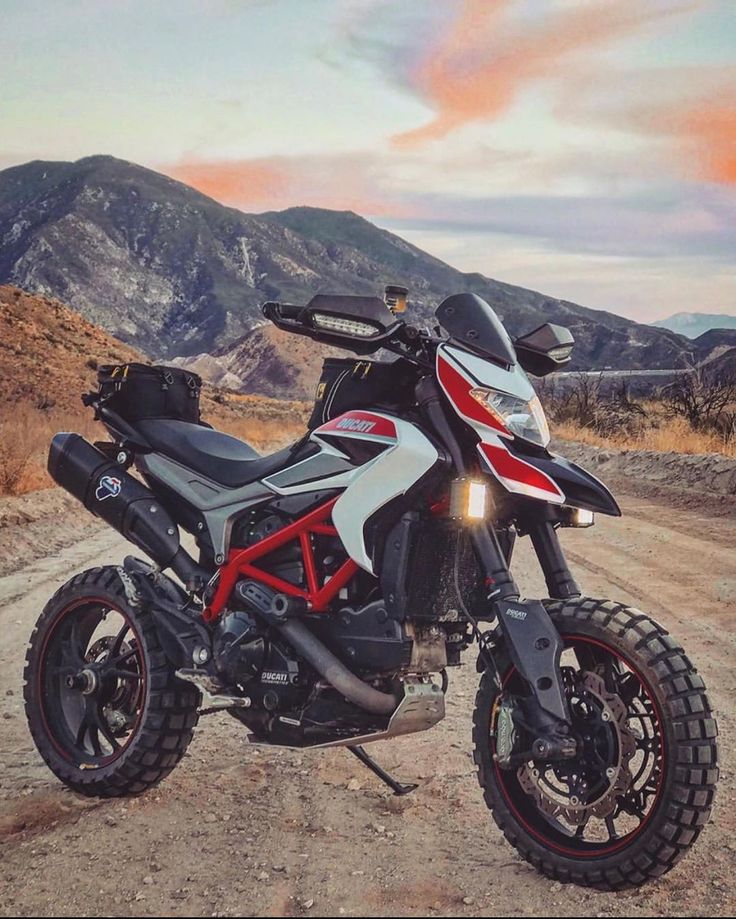 a red and white motorcycle parked on top of a dirt road next to mountains in the background