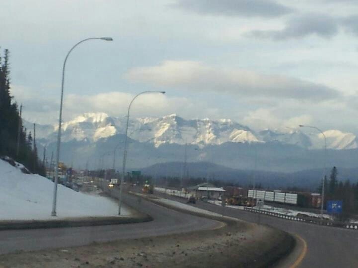 the mountains are covered in snow as cars drive down the road near them on a cloudy day