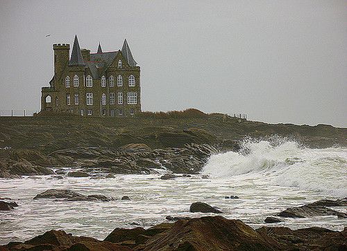 an old house sitting on top of a rocky cliff next to the ocean with waves crashing in front of it