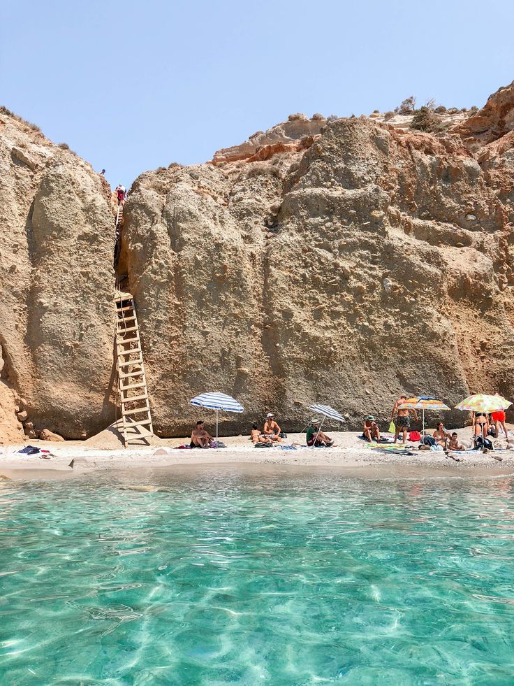 people are sitting on the beach under umbrellas near large rocks and clear blue water