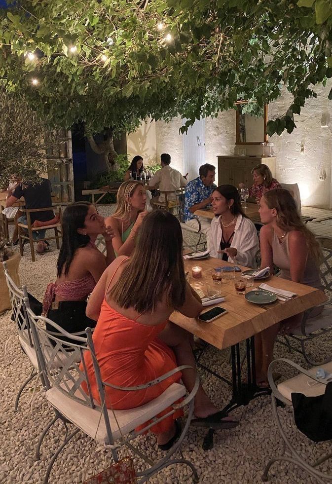 a group of people sitting around a table eating food under a tree with lights strung overhead