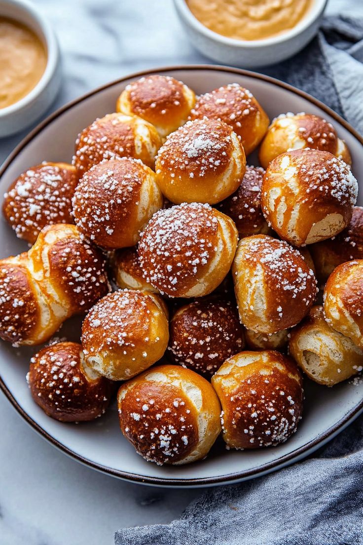 a white plate topped with mini pretzels covered in powdered sugar next to bowls of dipping sauce