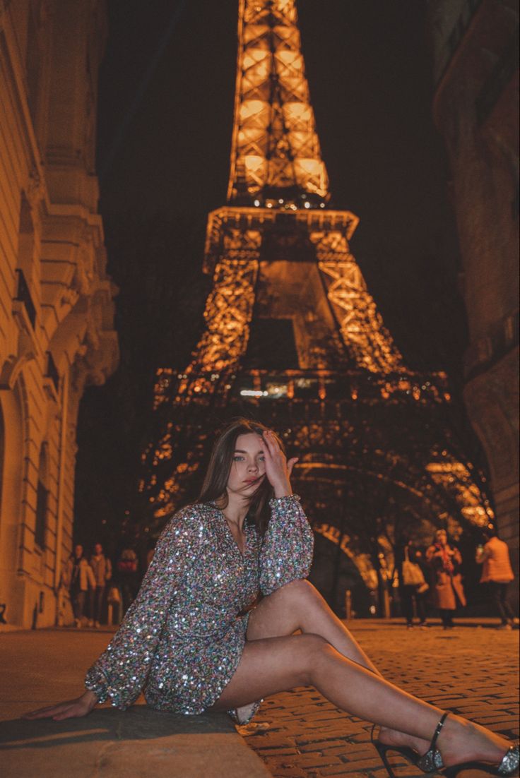 a woman sitting on the ground in front of the eiffel tower at night