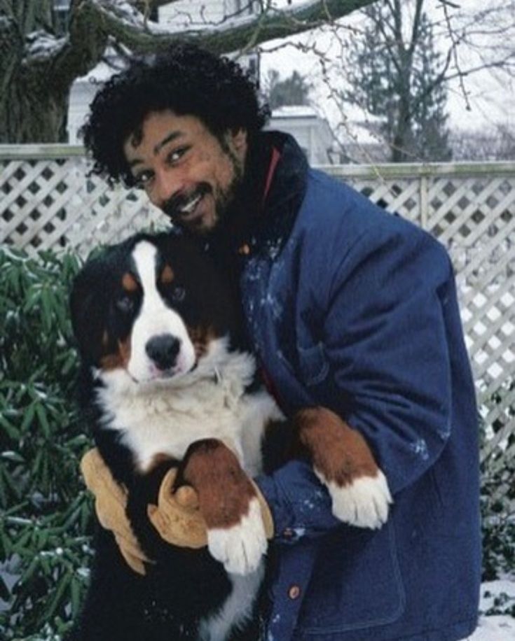 a man holding a dog in his arms while standing next to a bush covered with snow