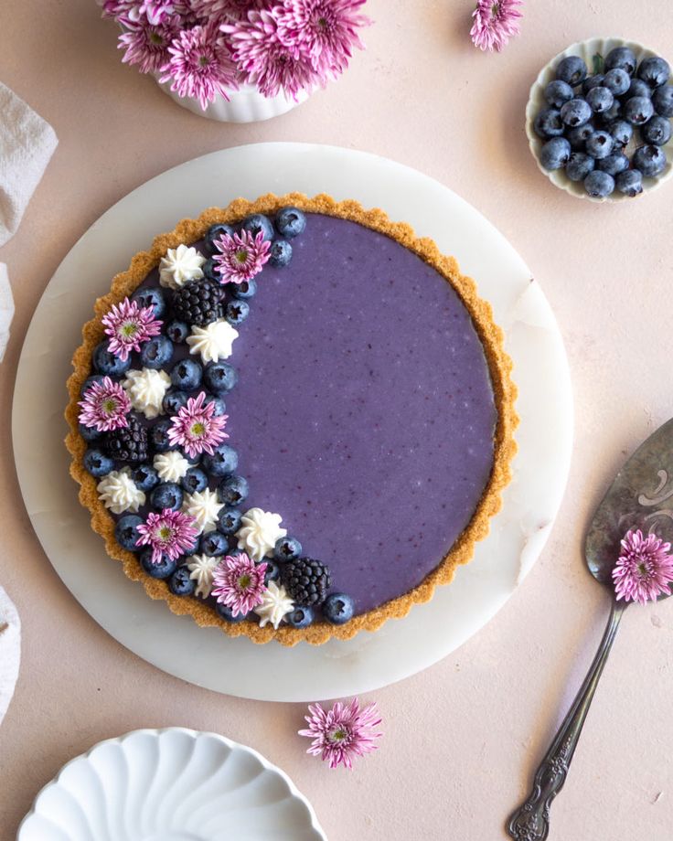 a blueberry cheesecake on a white plate with flowers and spoons next to it