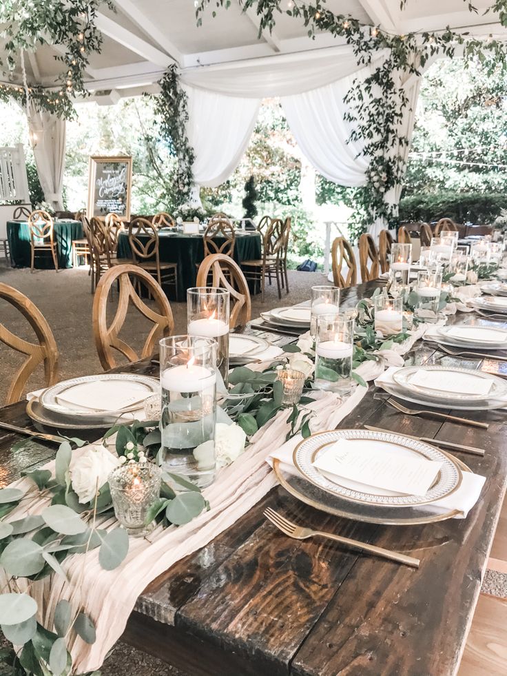 a table set up with place settings and greenery on the tables for an outdoor wedding reception