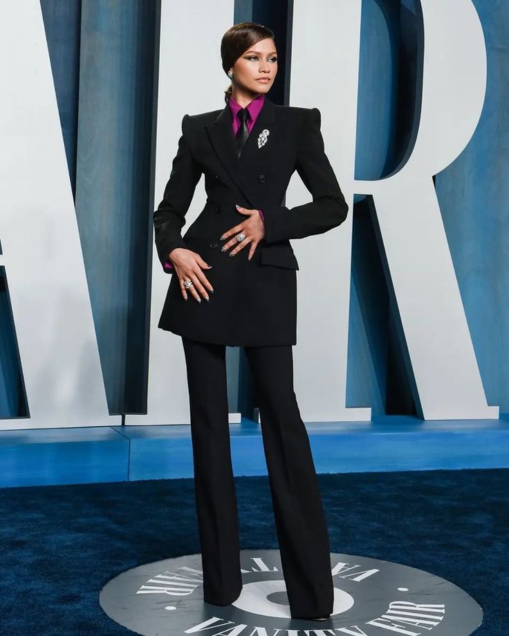 a woman in a black suit and pink shirt standing on a blue carpet with her hands on her hips