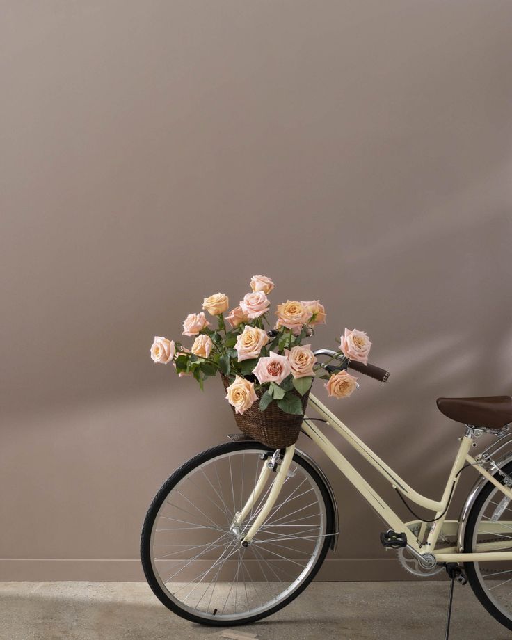 a bicycle with flowers in the basket parked next to a pink wall and wooden chair