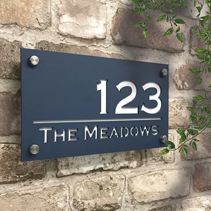 a metal house sign mounted to the side of a brick wall that reads, 123 the meadows