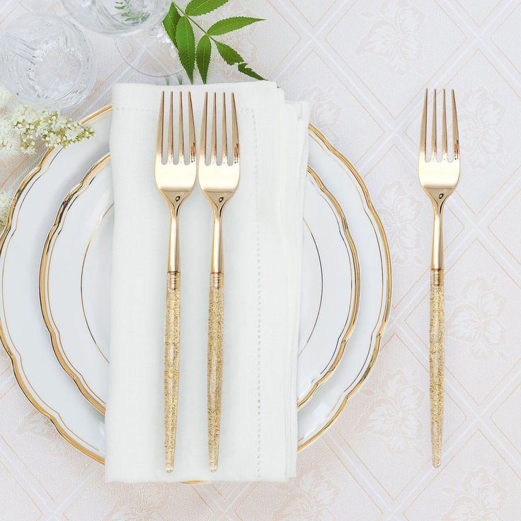 a white plate topped with two gold forks next to a vase filled with flowers and greenery