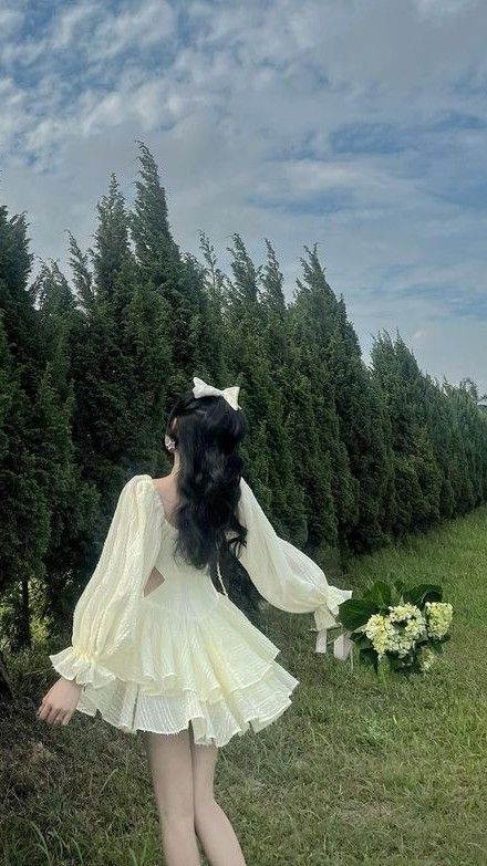 a woman with long black hair wearing a white dress standing in front of some trees