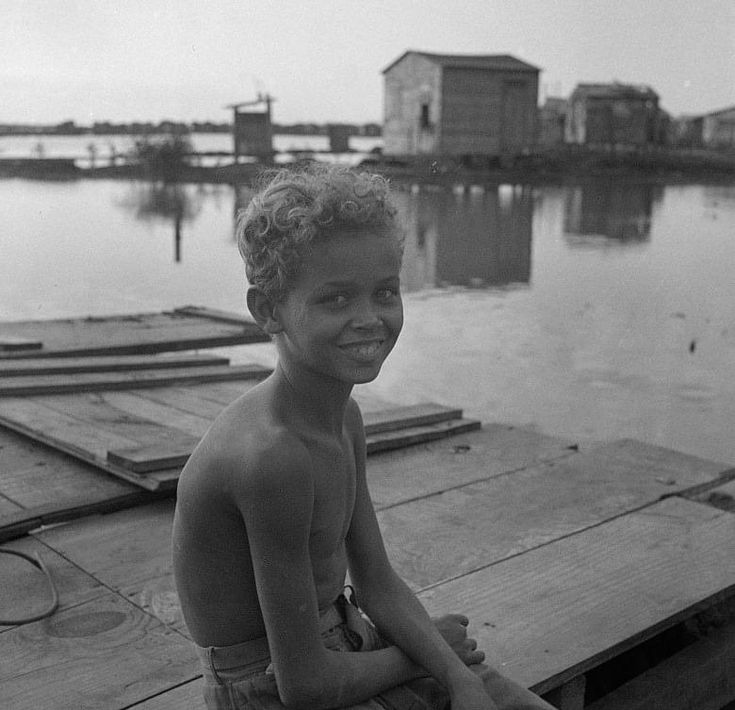 a young boy sitting on top of a wooden dock next to water and buildings in the background