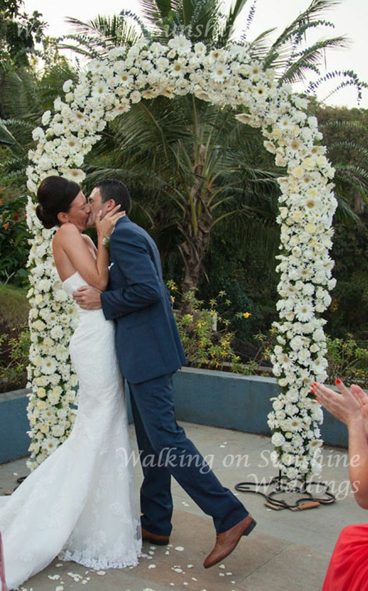 a bride and groom kissing in front of an arch with white flowers on the side