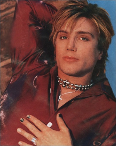 a man with his hand on his chest posing for a magazine cover, wearing a red leather jacket and beaded necklace