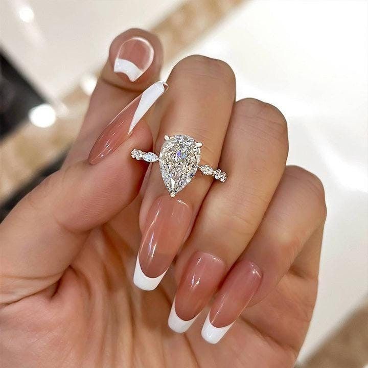 a woman's hand holding a ring with a diamond on it and white nails