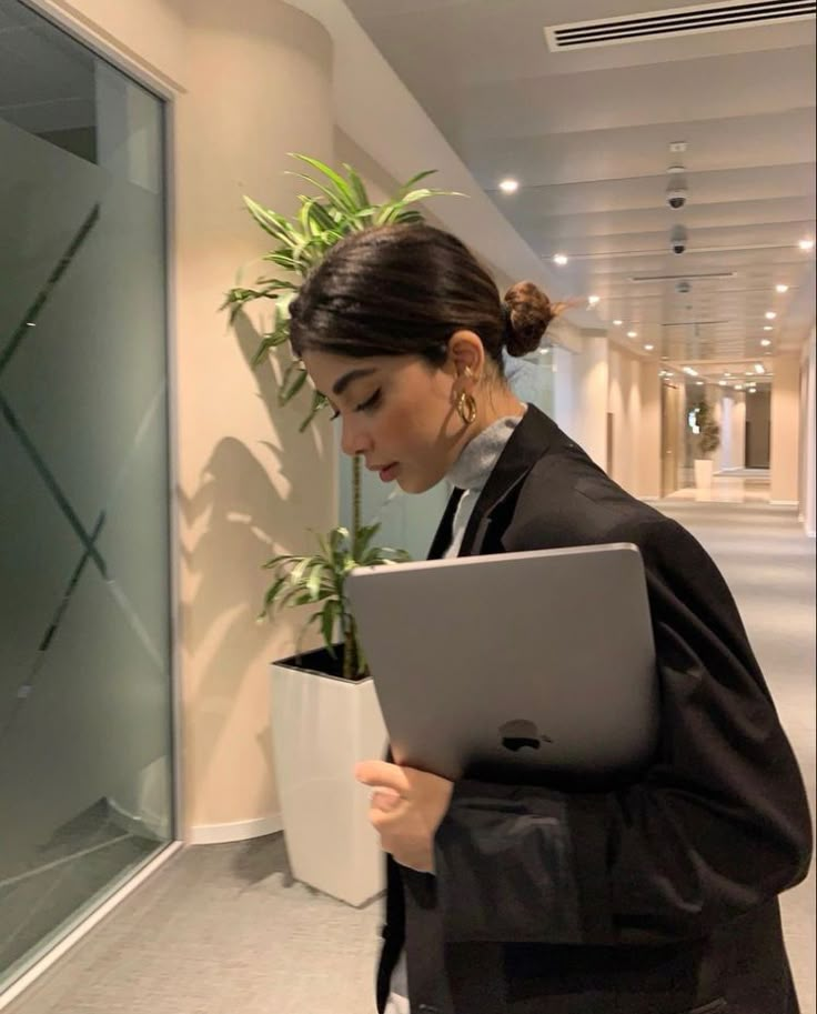 a woman standing in an office holding a laptop and looking down at the screen with her hands on her hips