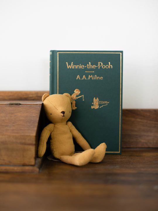 a brown teddy bear sitting next to a book on a wooden shelf in front of a white wall