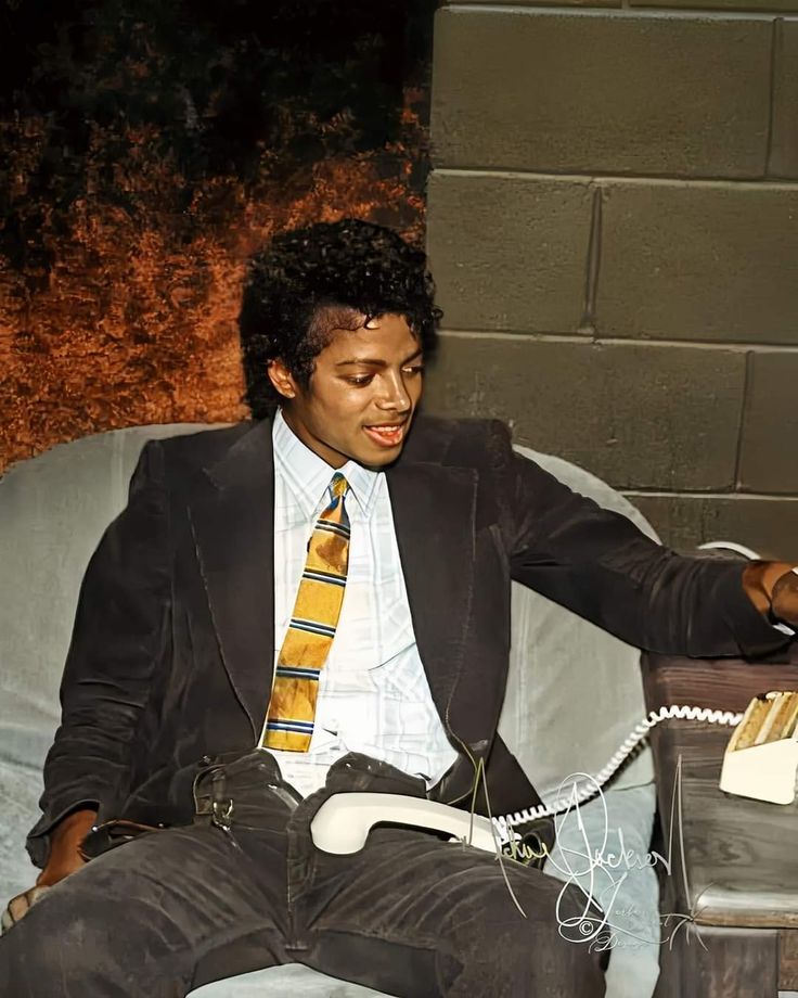 a man in a suit sitting on a couch talking on a phone with his hand