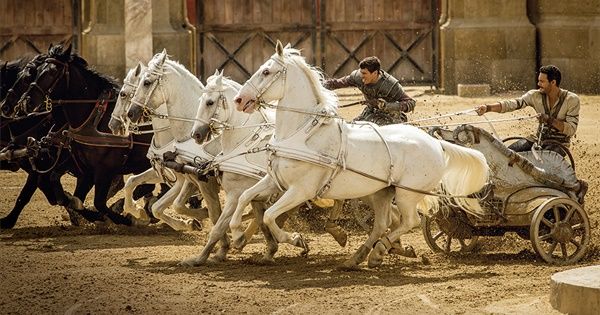 three horses pulling a man in a cart with two men on it and another horse behind them