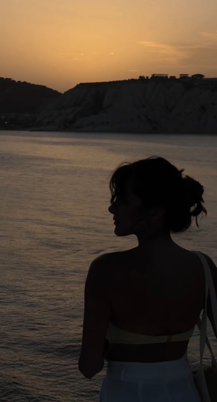 a woman standing in front of the ocean at sunset with her back to the camera