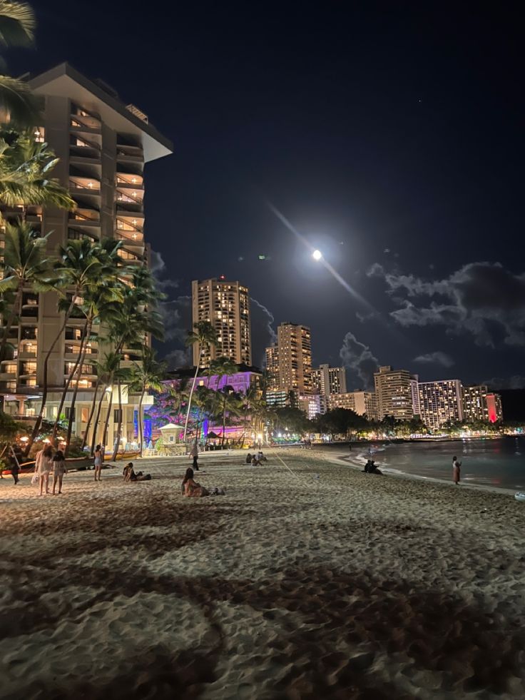 people are on the beach at night in front of some tall buildings and palm trees