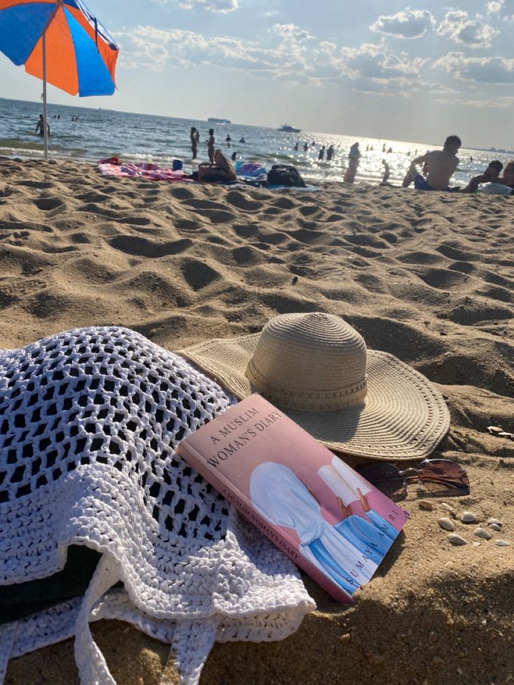 a book and hat on the beach with people in the water behind it, under an umbrella