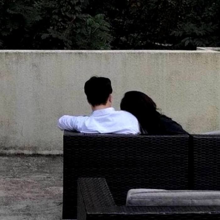 a man and woman sitting on top of a couch next to each other in front of a cement wall