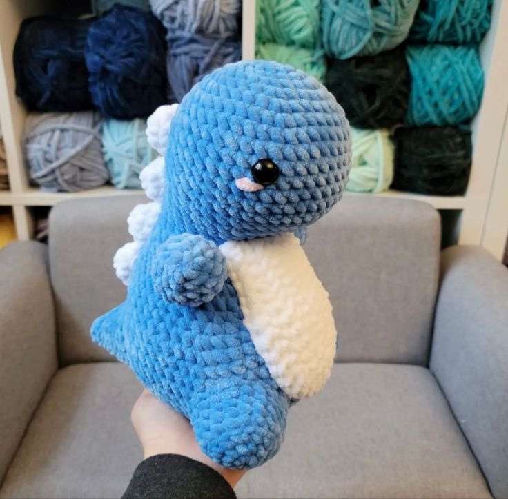 a hand holding a blue crocheted stuffed animal in front of a gray couch